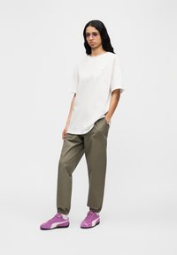 Γυναίκα που φοράει υπερμεγέθη λευκό μπλουζάκι, χακί παντελόνι φόρμας, μοβ αθλητικά παπούτσια και ορθογώνια γυαλιά ηλίου με απόχρωση, στέκεται μπροστά σε απλό φόντο.