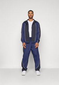 Marine tracksuit med en lynlåsåben jakke med hvide kanter og matchende bukser med snor i taljen. Materialet virker let og glat.