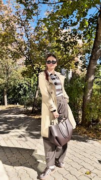 Beżowy trencz, pasiasta koszula z długim rękawem, brązowe spodnie z wysokim stanem, czarne okulary przeciwsłoneczne, elegancka brązowa torebka, sportowe czarne trampki, sceneria na zewnątrz.