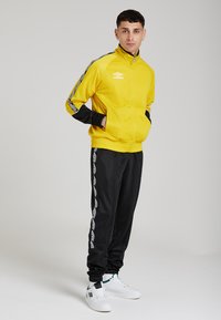 Geel trainingsjack met zwarte accenten, ritssluiting aan de voorkant en logo. Zwarte trainingsbroek met een gestreept patroon. Een paar witte sneakers.