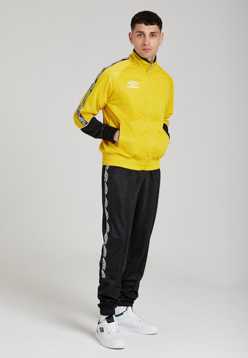Geel trainingsjack met zwarte accenten, ritssluiting aan de voorkant en logo. Zwarte trainingsbroek met een gestreept patroon. Een paar witte sneakers.