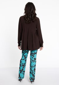 Braunes Langarmoberteil mit Falten im Rücken, kombiniert mit türkisfarbenen, floral gemusterten Hosen an einem Model, das schwarze Absätze trägt.