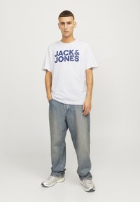 Tricou alb cu mânecă scurtă, inscripționat cu textul albastru "JACK & JONES", asortat cu blugi lejeri de culoare albastru deschis și pantofi sport argintii.