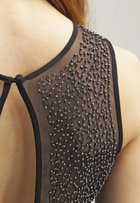 Černý síťový top s jemným korálkovým zdobením a kulatým výstřihem, s klíčovou dírkou na zadní straně a hladkou texturou látky.