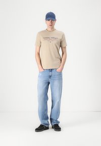 Beige bomull t-skjorte med "Tommy Hilfiger" logo, lyseblå slitte jeans, svarte sneakers og en blå caps.
