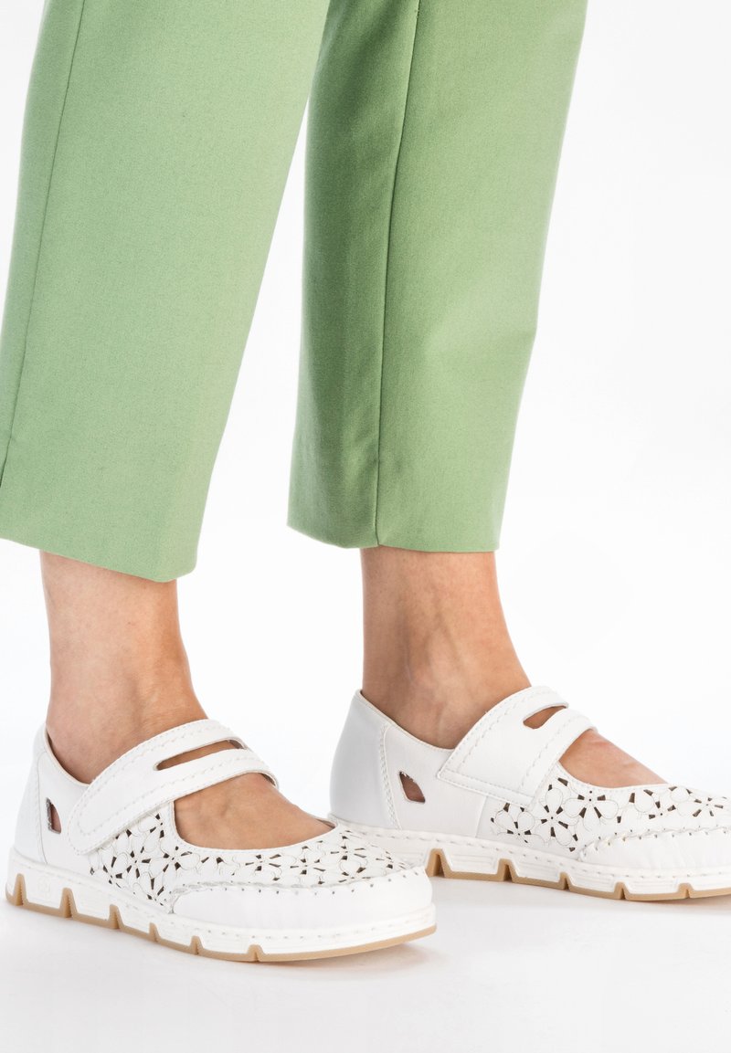 Chaussures blanches de style Mary Jane avec un design perforé et une sangle réglable ; semelle en caoutchouc ; associées à un pantalon court vert.