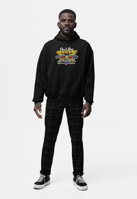 Uomo in piedi che indossa una felpa nera con cappuccio con una grafica colorata "Don't Stop Believin' Journey", pantaloni a scacchi neri e sneaker nere.