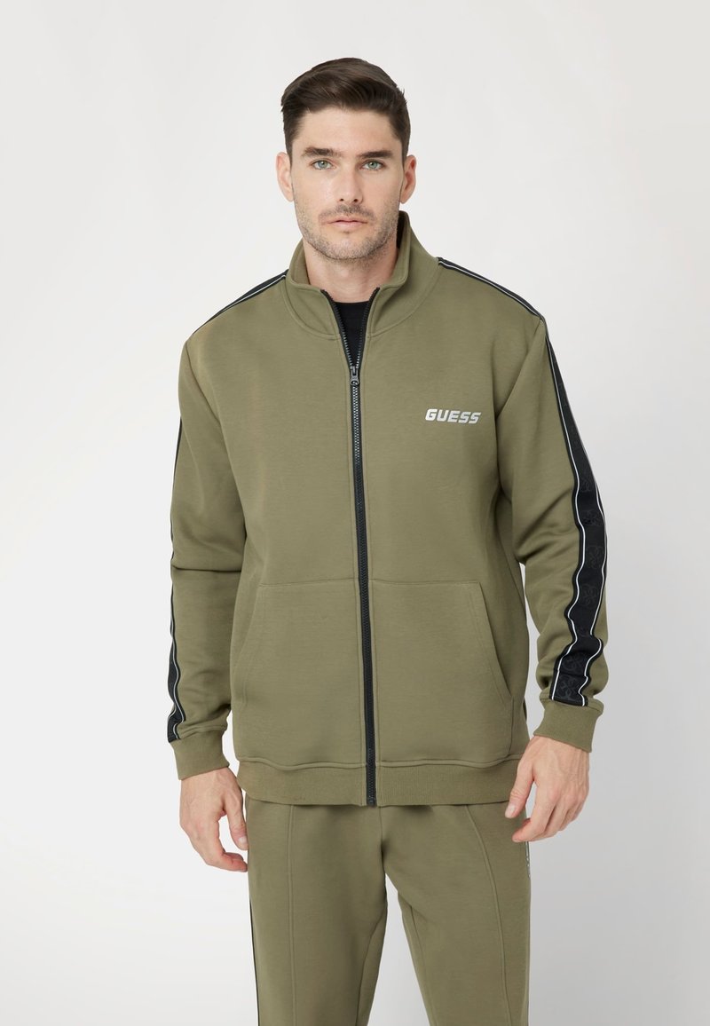 Giacca da tracksuit verde oliva con zip completa, colletto alto, strisce nere di accento e tasche frontali. Presenta il logo sul petto.