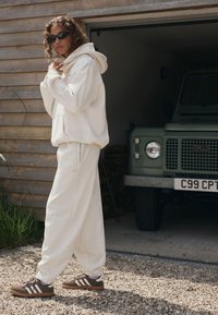 Balta laisvo stiliaus džemperis su gaubtu ir derančios laisvalaikio kelnės iš minkštos medžiagos. Rudi sportbačiai su trim baltomis juostelėmis. Fone matomas žalias automobilis.