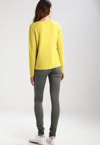 Gelbes Strickpullover mit Rundhalsausschnitt, strukturiertem Muster und seitlichen Nähten, kombiniert mit grauen Skinny-Jeans und weißen Sneakers.