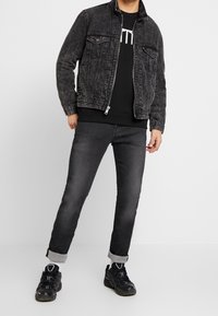 Hombre vistiendo una chaqueta de mezclilla negra con efecto de lavado ácido, sudadera negra con estampado blanco, jeans oscuros con las piernas remangadas, calcetines blancos y zapatillas negras, de pie sobre un suelo blanco.