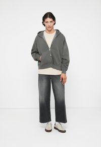 Harmaa hupparimekko vetoketjulla ja etutaskuilla, yhdistettynä vaalean beigeen t-paitaan ja leveisiin harmaisiin farkkuihin. Valkoiset tennarit ruskeilla pohjilla.