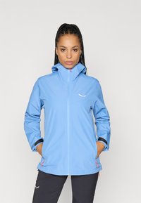 Veste imperméable bleu clair avec un col haut, une fermeture éclair à l'avant et des poignets élastiques. Présente un logo sur la poitrine et des accents zippés orange.