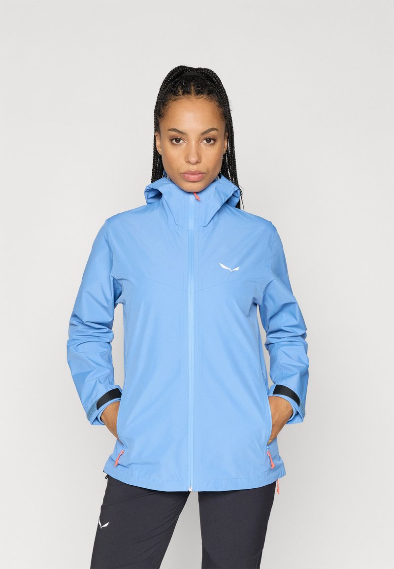 Veste imperméable bleu clair avec un col haut, une fermeture éclair à l'avant et des poignets élastiques. Présente un logo sur la poitrine et des accents zippés orange.