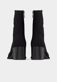 Ζευγάρι μαύρα μποτάκια αστραγάλου με ψηλό τακούνι και φαρδιά τακούνια, με φερμουάρ στο πίσω μέρος, τοποθετημένα πάνω σε απλό λευκό φόντο.