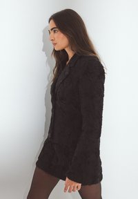 Mustarikk mustraseelne bleiser, millel on vormitud lõige, lõigatud krae ja pikad varrukad, koos musta mini seelikuga ja sukkpükstega.