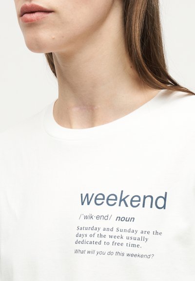 Νεαρή γυναίκα φορώντας ένα λευκό t-shirt με τη λέξη «weekend» και τον ορισμό της τυπωμένο στο μπροστινό μέρος.