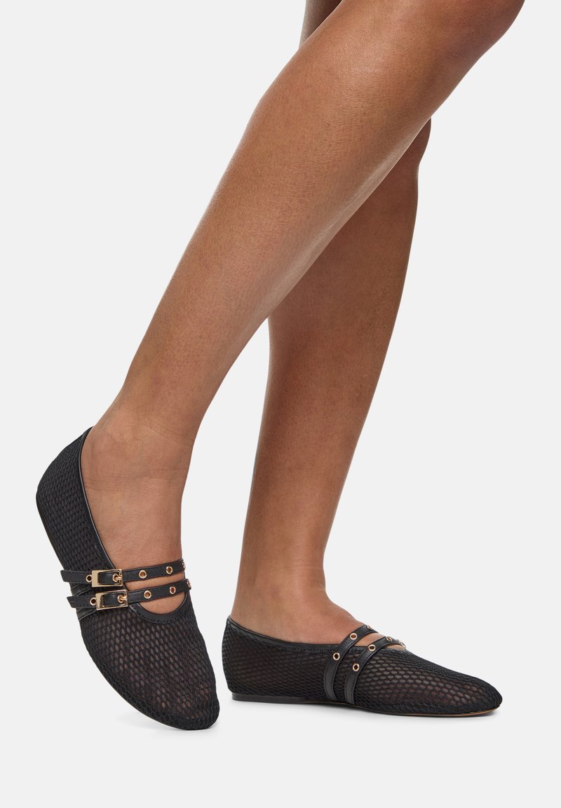 Gambe femminili che indossano ballerine nere in mesh con doppie cinghie e dettagli in oro su uno sfondo bianco.