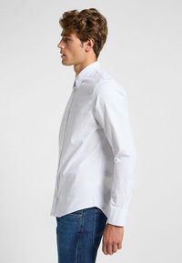 Camisa de botón azul claro en una mezcla de algodón, con un cuello puntiagudo, mangas largas y un dobladillo ligeramente curvado. Sin patrones visibles.