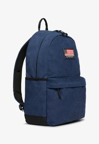 Mochila de tela azul con bolsillo frontal con cremallera, base negra, correas acolchadas y un parche con la bandera estadounidense y el texto "Superdry".
