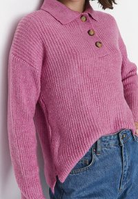 Eine Frau trägt einen rosa gestrickten Pullover mit drei braunen Knöpfen und blaue Denim-Jeans und steht vor einem weißen Hintergrund.