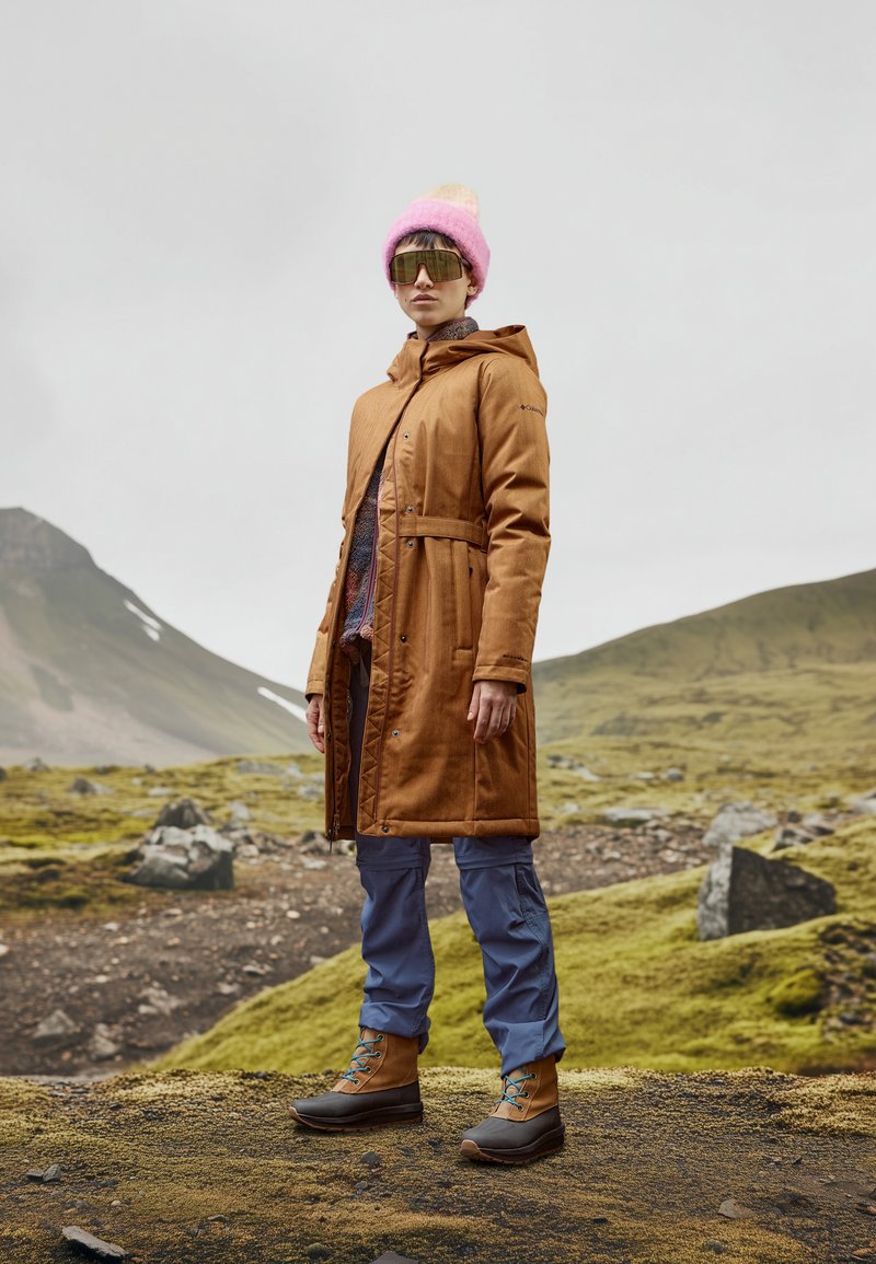 Brauner wasserdichter Mantel mit einem Knotenbelt, graue und braune Stiefel mit blauen Schnürsenkeln, rosa Strickmütze und graue Sonnenbrille. Bergige Kulisse.
