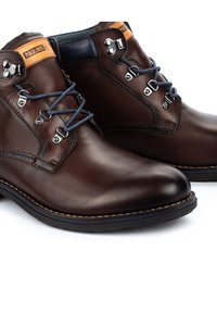 Bottes en cuir marron à la cheville avec des accents bleu foncé, dotées d'œillets en métal et de lacets marins. Texture lisse avec des détails de couture contrastants.