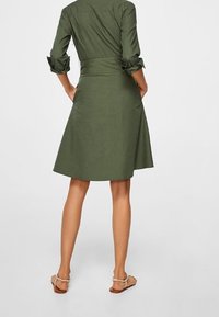 Femme portant une robe olive à mi-genoux avec des manches retroussées et des sandales beiges, se tenant les mains dans les poches arrière contre un fond uni.