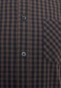 Chemise à carreaux marron et noire boutonnée avec poche avant présentant un motif diagonal opposé au tissu principal.