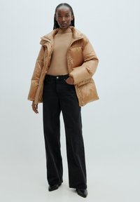 Puffjacke in glänzendem Karamell mit großem Kragen, kombiniert mit einem figurbetonten beigen Oberteil und hoch taillierten schwarzen Jeans. Schwarze spitze Schuhe.