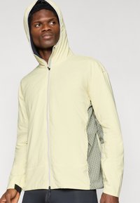 Světle žlutá zipová bunda s kapucí, zahrnující texturovaný olivově zelený boční panel a elastické manžety pro pohodlné nošení.