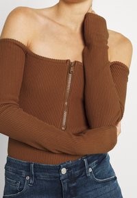Hnědý žebrovaný body s off-shoulder designem a předním zipem. Dlouhé rukávy sahají až na zápěstí, doplněné tmavě modrými džíny.