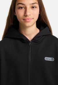 Giovane con lunghi capelli castani che indossa una felpa nera con zip e un piccolo logo "element" sul lato sinistro del petto, su uno sfondo neutro.