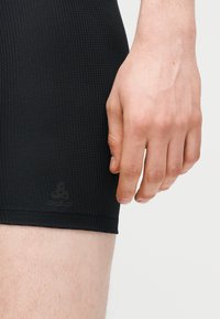 Main droite reposant à côté de la cuisse, portant un short moulant noir texturé avec un logo de marque discret près de l'ourlet.