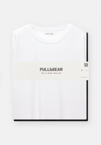 T-shirt en coton blanc soigneusement plié, avec une large bordure noire en bas et une étiquette avec "PULL&BEAR" et du texte en dessous.