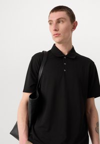 Černé polo tričko s jemným vertikálním proužkovým vzorem, krátkými rukávy, tříknoflíkovým zapínáním a měkkou texturou. V kombinaci s černou koženou taškou.