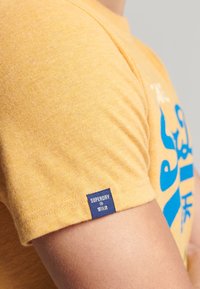Camiseta de algodón amarilla con un estampado gráfico azul en la parte delantera, que presenta una pequeña etiqueta navy con "SUPERDRY" en la manga.