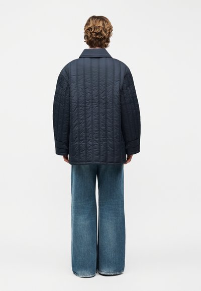 Άτομο με κοντά σγουρά μαλλιά που φοράει μαύρο μπουφάν με επένδυση και φαρδιά μπλε τζιν, στέκεται γυρισμένο ανάποδα μπροστά σε λευκό απλό φόντο.