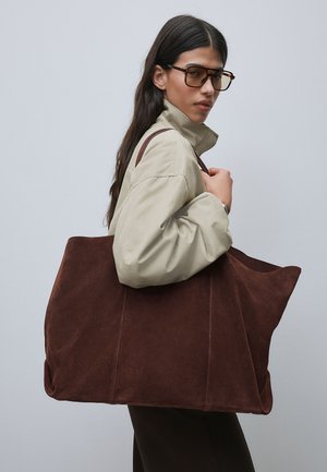 Kvinde med langt mørkt hår iført beige jakke og store tonede briller, bærer en stor mørkebrun ruskindstaske på skulderen.