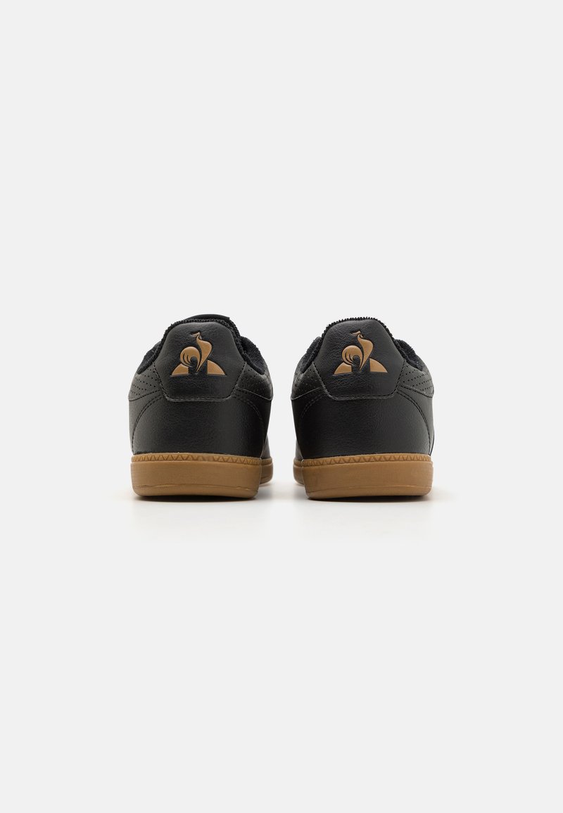 Zapatillas de cuero negro con un acabado texturizado, que cuentan con una suela de goma y un emblema dorado en la parte posterior. Diseño de corte bajo.