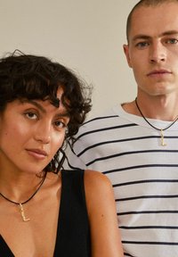 Guld berlockhalsband med en liten "l"-form på ett svart snöre. Modellen bär en svart ärmlös topp; den andra modellen bär en randig skjorta.