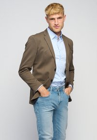 Brun fitted blazer over en lys blå stribet skjorte, parret med lyseblå jeans. Blazeren har to knapper og sidelommer.