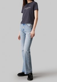 Camiseta corta de color gris oscuro con texto blanco, combinada con jeans acampanados de color azul claro. Modelo vistiendo zapatos negros, de pie frente a un fondo neutro.
