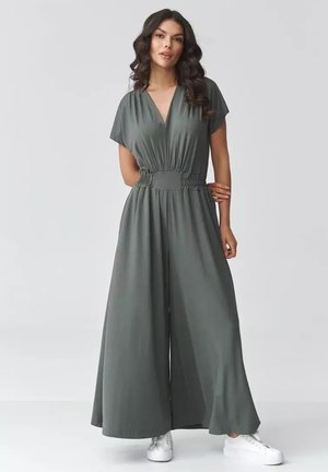 Kobieta o długich ciemnych włosach, ubrana w szary kombinezon z szerokimi nogawkami i białe trampki, stoi z jedną ręką na biodrze.