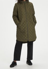 Manteau long matelassé vert olive avec capuche, doté d'une fermeture éclair à l'avant, de poches latérales et de boutons-pression sur l'ourlet. Pantalon et chaussures noirs.