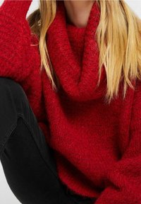 Blonde Person, der einen dicken, klobigen roten Strickpullover mit großem Kragen trägt, kombiniert mit schwarzen Hosen, sitzt mit einem Knie angezogen.