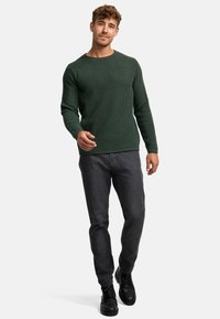Pull en maille côtelée vert foncé avec un col rond, associé à un jean gris foncé et des bottes noires. Le modèle se tient devant un fond neutre.