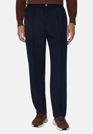 Uomo che indossa pantaloni blu navy con pieghe frontali e scarpe da ginnastica marroni su sfondo chiaro.
