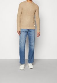 Beige neulepaita, jossa on teksturoitu kuvio, pyöreä pääntie ja pitkät hihat, yhdistettynä vaaleansinisiin suoriin farkkuihin ja valkoisiin lenkkareihin.