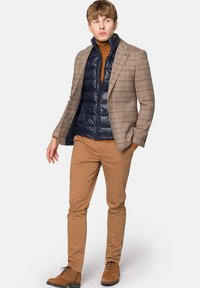 Męski zestaw ubrań z brązową w kratę marynarką na granatowej kamizelce puchowej, beżowymi spodniami i brązowymi butami na sznurówki. Gładkie tekstury materiałów i kontrastujące kolory.